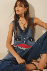 Chaleco de denim rígido azul con efecto desgastado, corte ajustado y escote en V. Presenta botones metálicos grabados en el frente, dos bolsillos con tapa en el pecho y un detalle de tira de ajuste en la espalda.