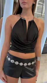 Top negro de satén, sin mangas, con escote halter adornado con un detalle metálico rígido alrededor del cuello y abertura frontal en V. Presenta diseño drapeado en el delantero y corte ajustado.