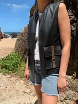 Chaleco negro de cuero con cuello redondo y cierre frontal. Presenta bolsillos utilitarios con solapa y detalles en cuero marrón en los cierres de los bolsillos.