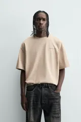 Camiseta de manga corta y cuello redondo, color beige claro, con corte relajado (relaxed fit) y un pequeño texto bordado en el pecho.