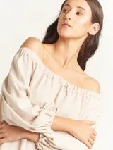 Vestido largo color beige, con escote elastizado que permite usarlo con los hombros descubiertos, mangas largas abullonadas, bolsillos laterales y tajos laterales en el ruedo.