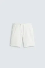 Bermuda blanca de tela lisa, con cintura elástica ajustable mediante cordón blanco. Presenta pliegues frontales, bolsillos laterales y bolsillos de vivo en la espalda. El bajo tiene un dobladillo vuelto.