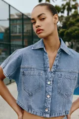 Camisa corta de denim en color azul claro, con cuello solapa, escote en V y manga corta. Presenta bolsillos delanteros de parche con solapa y cierre frontal de botones metálicos.