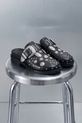 Mocasines negros de cuero sintético estilo zueco, con hebilla lateral plateada y apliques metálicos estilo western en toda la capellada.