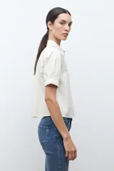 Camisa de algodón color beige claro, con cuello camisero, botones frontales y mangas cortas abullonadas con elástico en el puño.