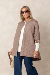 Campera acolchada de corte amplio y largo midi, color beige con diseño capitoneado. Presenta mangas tres cuartos con puños vueltos que revelan un interior estampado con motivo animal print. Diseño sin cuello y cierre frontal oculto.