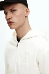 Campera con capucha de algodón pesado, color blanco hueso, de corte holgado y hombros caídos. Posee cierre frontal bidireccional, bolsillos canguro delanteros y puños y bajo acanalados.