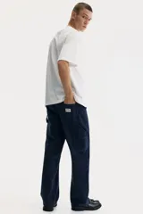 Pantalón tipo worker confeccionado en sarga de algodón de color azul marino, con un corte relaxed fit y piernas rectas. Presenta una cintura clásica con cierre de botón, bolsillos laterales al bies, bolsillos traseros, una trabilla funcional en un lateral y un bolsillo cargo dividido en el otro.
