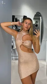 Vestido corto ajustado color oro rosado con acabado brillante. Diseño asimétrico con cuello alto y un gran recorte lateral que expone el abdomen, unido por un detalle fruncido con argolla.