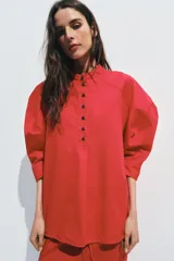 Camisa de algodón con cuello redondo y cierre frontal de botones a contraste. Presenta mangas abullonadas por debajo del codo y bajo asimétrico con aberturas laterales.