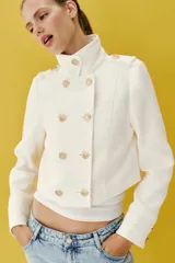 Chaqueta corta blanca de estilo militar, con cuello subido, manga larga y trabillas desflecadas en los hombros. Cuenta con cierre frontal cruzado decorado con botones metálicos dorados.
