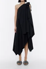 Vestido midi negro asimétrico con un solo hombro y lazo decorativo. Presenta múltiples capas y un corte irregular en el ruedo.