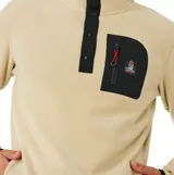 Buzo de polar beige con cuello alto y cierre frontal parcial de botones a presión. Presenta un bolsillo frontal en el pecho con cierre y detalles en negro en el cuello, bolsillo y puños.