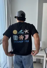 Remera negra de manga corta con estampado gráfico en la espalda que incluye un sol amarillo, una estrella de mar, una flor, una ola con el logo 'Brokers' y formas abstractas en tonos pastel, junto al texto 'VITAMIN SEA'.