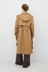 Trench coat color beige con cuello de solapa, cierre frontal abotonado, bolsillos laterales y mangas largas con cintas ajustables. Incluye capucha desmontable y forro interior rayado. La tela exterior cuenta con protección repelente a la lluvia y manchas.