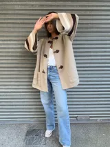 Saco color beige de lana con cierre frontal con broches tipo tanca de cuero. Presenta mangas anchas con detalle de rayas horizontales en marrón y beige.