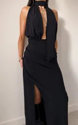 Vestido largo negro con escote halter profundo y detalle de lazo al cuello. Presenta un diseño drapeado en la cintura y una abertura lateral pronunciada en la falda.