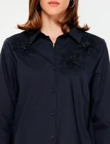 Camisa blanca de manga larga con cuello clásico y cierre frontal de botones. Presenta detalles bordados florales tono sobre tono en la zona de los hombros y el pecho.