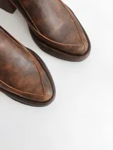 Zapato tipo zueco de cuero color chocolate con efecto desgastado, plataforma y taco cuadrado. Presenta una tira trasera ajustable con hebilla metálica.