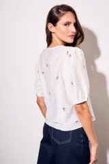 Blusa de viscosa color marrón, con escote en V, mangas cortas abullonadas y cierre frontal con botones. Cuenta con pequeños detalles bordados distribuidos por la prenda.