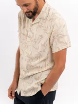 Camisa de manga corta color beige con estampado tropical de hojas, cuello camisero abierto y cierre frontal de botones.