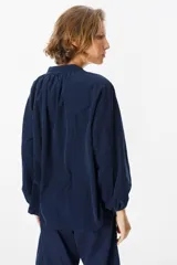 Camisa de pana color verde oliva, con cuello mao, cierre frontal de botones y mangas largas abullonadas con puños elásticos.