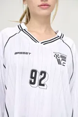 Camiseta blanca de manga larga con cuello en V y detalles en negro. Presenta el logo de la marca en el pecho, el número 92 y un escudo con tres balones de fútbol.
