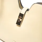 Cartera de mano estructurada color beige, con asa corta fija y correa larga regulable y desmontable. Presenta un cierre superior con broche giratorio metálico dorado y logo de la marca aplicado en el frente.
