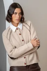 Saco de punto color beige, con cierre frontal de botones marrones y detalle de costuras decorativas oscuras en los hombros. Presenta un diseño clásico con cuello redondo.