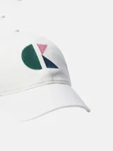 Gorra de seis paneles color blanco, con visera curva y cierre ajustable. Presenta un monograma bordado en el frente compuesto por formas geométricas en verde oscuro, rosa pálido y azul oscuro.