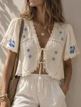 Camisa corta de lino color crudo, con escote en V, mangas cortas abullonadas y cierre frontal con lazos. Presenta bordados florales en tonos azules y detalles de encaje en los bordes.
