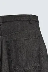 Pantalón de jeans de algodón ligero, color negro lavado, con corte balloon fit (holgado y ancho). Presenta pliegues frontales marcados en la cintura y cierre frontal de cremallera y botón.