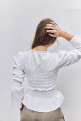 Blusa blanca de algodón con cuerpo fruncido y elástico, cuello redondo con detalle de puntilla bordada, mangas largas abullonadas con puños elásticos y ruedo con volado.