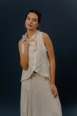 Chaleco de lino color marfil o beige claro, con diseño sin mangas, cuello alto amplio, cierre frontal con botones y bolsillos delanteros amplios. Presenta un corte asimétrico y relajado.