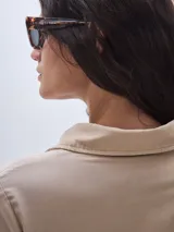 Vestido camisero largo color beige, con cuello clásico, mangas largas y cierre frontal con botones. Presenta un nudo decorativo a la altura de la cintura y una abertura en el ruedo.