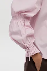 Camisa blanca de manga larga abullonada con detalle de punto smock en los puños y cordones para atar. Presenta cuello clásico y cierre frontal de botones.
