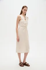Vestido midi de viscosa color beige, sin mangas y con cuello a la base. Presenta una abertura frontal con botones decorativos y detalles de frunces en el centro del pecho.