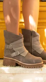 Botas cortas de estilo urbano, confeccionadas en una combinación de gamuza y cuero vacuno en tonos tierra. Presentan un diseño con tiras decorativas cruzadas en el empeine, hebillas metálicas ajustables, cierre lateral y suela de goma con plataforma dentada.