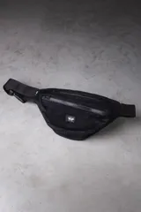 Riñonera de corderoy color negro, con compartimento principal y bolsillo frontal con cierre, tira ajustable con hebilla plástica y detalle de parche bordado con la marca VCP.