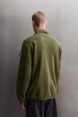 Chaqueta verde militar de punto con interior de polar. Tiene cuello alto, manga larga con bolsillo con cierre en la manga, bolsillos laterales con cierre y cierre frontal con cremallera.
