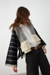 Chaqueta abierta de tejido de punto con diseño geométrico en blanco y negro. Presenta solapas con patrón de espiga y terminaciones con flecos largos en el frente. Posee mangas anchas con detalle de líneas horizontales punteadas.