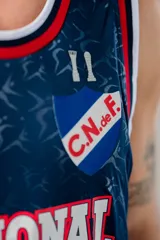 Musculosa de básquet Umbro del Club Nacional de Football, modelo alternativa 2025. Diseño en color azul marino con patrón sutil estampado, cuello redondo y sisas en rib con líneas blancas y rojas. Incluye el logo de Umbro, el escudo del club y la inscripción "Nacional" en el frente.