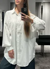 Camisa blanca de corte oversize, con cuello clásico, cierre frontal de botones y bolsillo en el pecho. Presenta mangas largas con puños abotonados y un diseño holgado con ruedo redondeado.