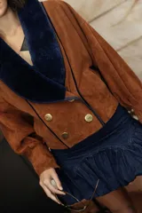 Chaqueta confeccionada en gamuza color negro con amplio cuello smoking de corderito en tono crudo. Presenta un diseño corto, cruzado con doble abotonadura y botones metálicos, mangas amplias y bolsillos.