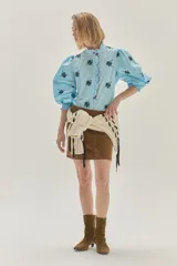 Blusa de algodón en color celeste con estampado floral en negro. Presenta cuello alto con volados y borde en color rojo, mangas abullonadas con puños elásticos tipo smock y cierre frontal con botones.