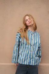 Camisa de denim azul con cuello mao, diseño holgado, mangas largas y cierre frontal con botones metálicos. Cuenta con costuras decorativas pespunteadas en el delantero.