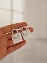 Par de aros colgantes con diseño geométrico compuesto por una pieza superior metálica de forma irregular y una pieza inferior cuadrada de resina con efecto nacarado.