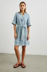 Vestido camisero corto de denim celeste con cuello en V, mangas cortas y cinturón de lazo. Presenta bolsillos laterales y pespuntes a contratono.