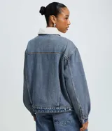 Campera trucker de jean azul con cuello de corderito blanco, cierre frontal con botones metálicos, bolsillos frontales con solapa y puños abotonados.