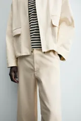 Pantalón chino de algodón color beige, de corte relajado (relaxed fit) y tiro medio. Presenta bolsillos delanteros y bolsillos traseros de vivo con cierre de cremallera y botón.